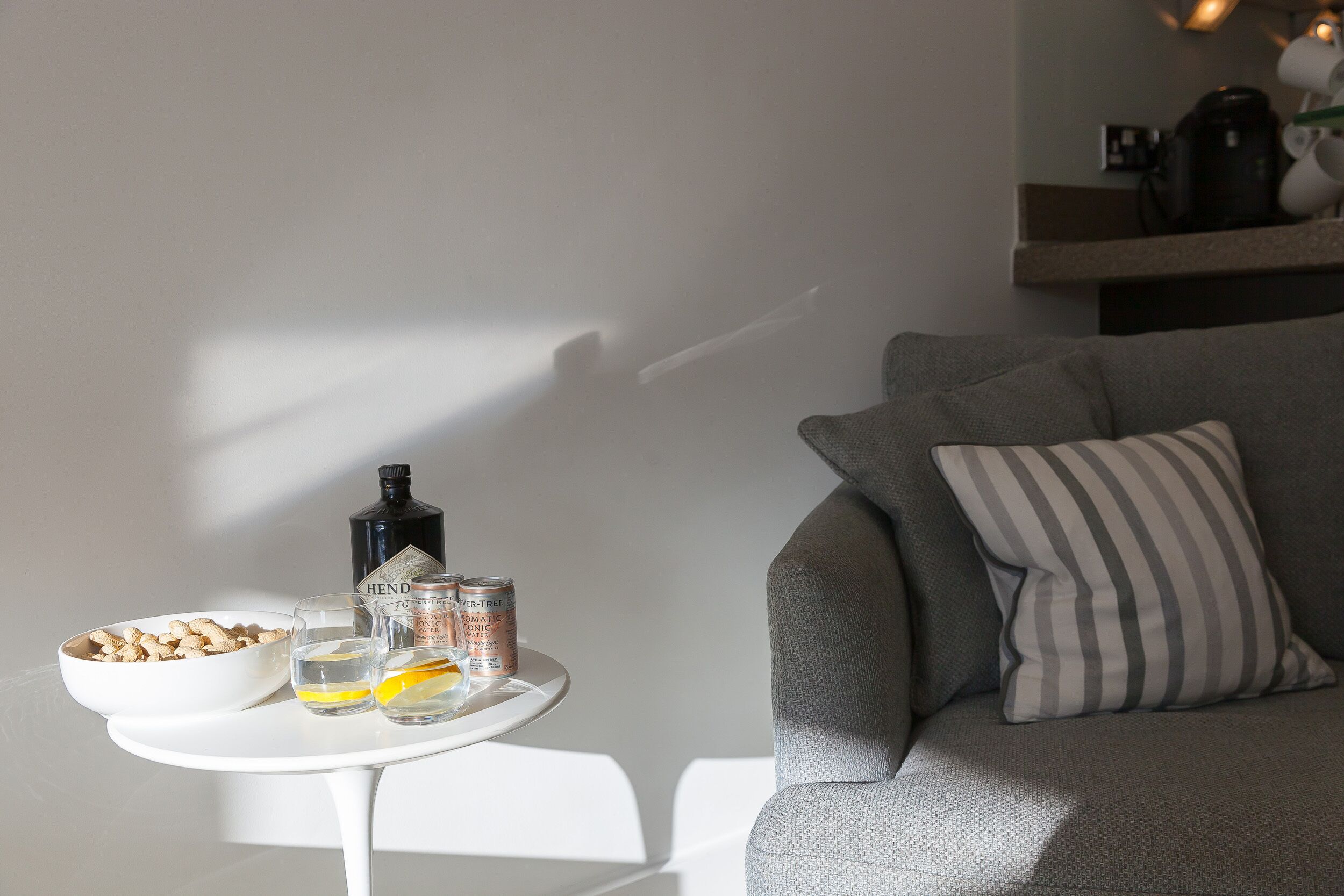 A modern living room with a grey sofa, a striped pillow, a small white table with a bowl of snacks, glasses, and a bottle.