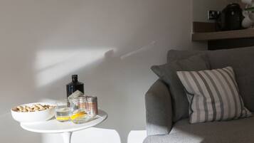 A modern living room with a grey sofa, a striped pillow, a small white table with a bowl of snacks, glasses, and a bottle.