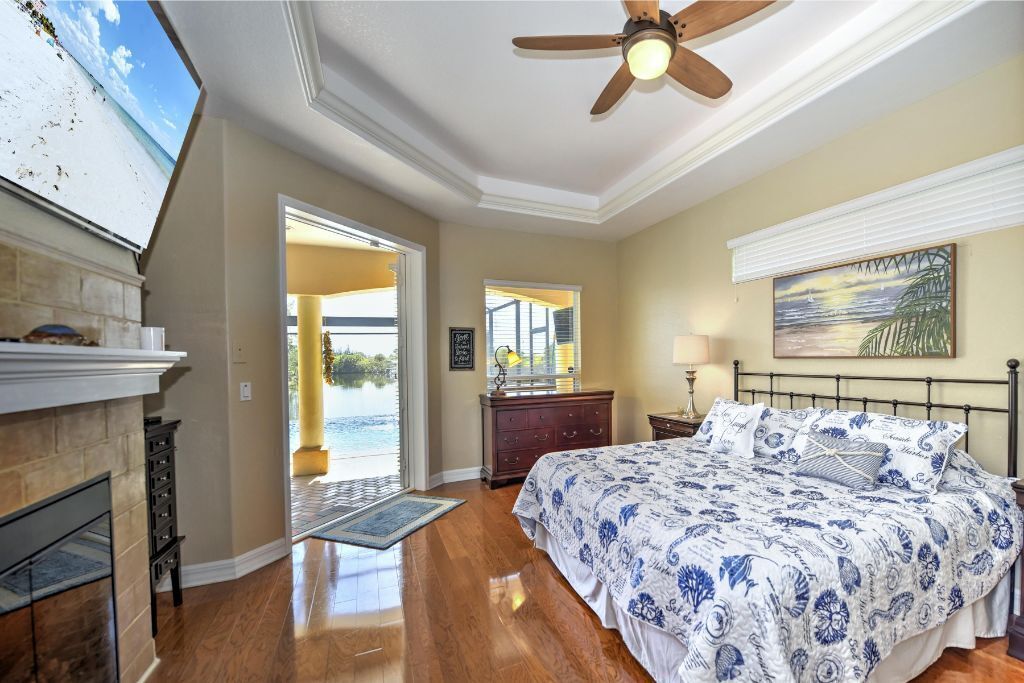 A bedroom with a large bed, a ceiling fan, a fireplace, and a view of the outdoors.