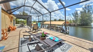 Un espacio al aire libre techado con vistas a un lago, amoblado con sillones de descanso y una mesa, y un cerramiento de vidrio.