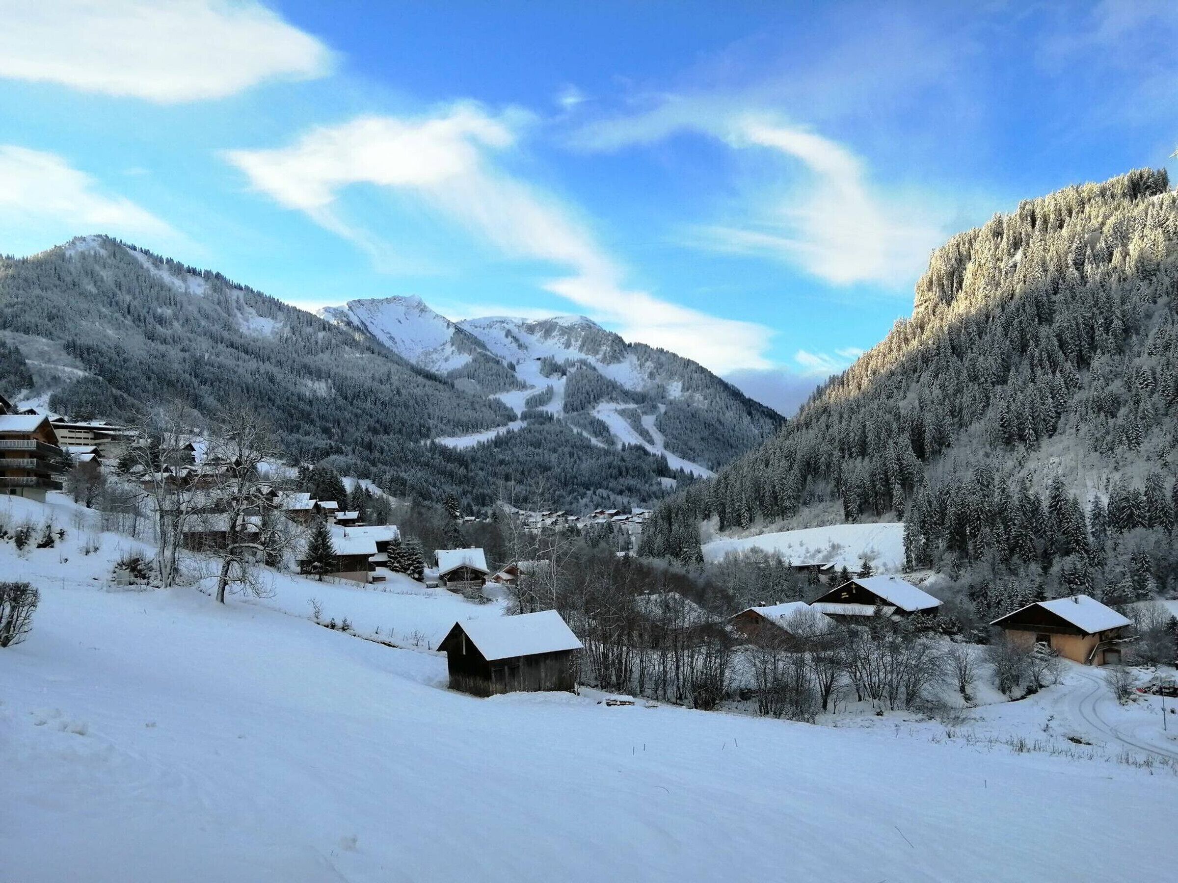 Uma aldeia montanhosa coberta de neve, com casas de madeira e uma pista de esqui ao fundo.