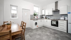 Een moderne keuken met witte kastjes, een ingebouwde oven, magnetron en wasmachine. Er staat een houten eettafel met stoelen en een raam zorgt voor natuurlijk licht.
