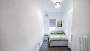 Een smalle gang die leidt naar een slaapkamer met een bed, nachtkastje, raam en een radiator.