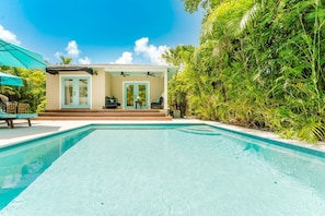 Una piscina con una veranda coperta, scale che conducono a una porta e rigogliosa vegetazione tutt'intorno alla proprietà.