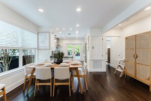 A modern dining area with a wooden table set for six, white chairs, and a view of the outdoors through large windows.