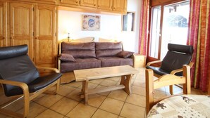 A living room with wooden cabinets, a brown sofa, two chairs, a wooden table, and a window with curtains.