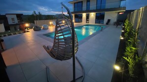 Un espace piscine extérieur moderne, doté d’un fauteuil suspendu, de chaises longues et d’un bâtiment bien éclairé en arrière-plan.