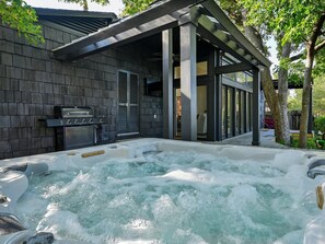 Relax in the hot tub, surrounded by a pergola and trees, with a grill nearby.