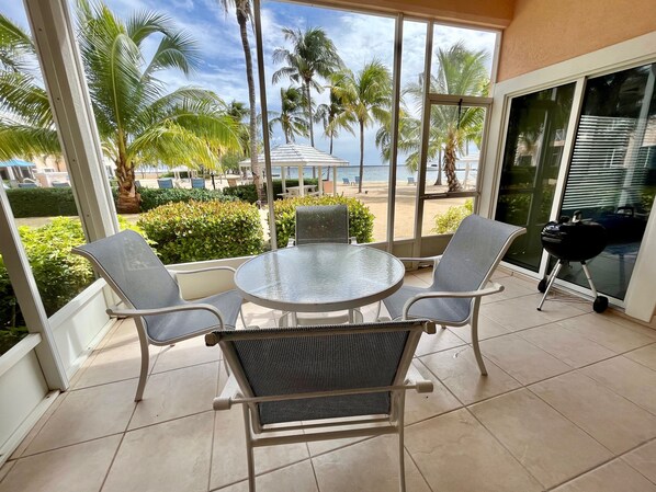 A balcony with a glass table and two chairs, overlooking a beach with palm trees and a clear sky.