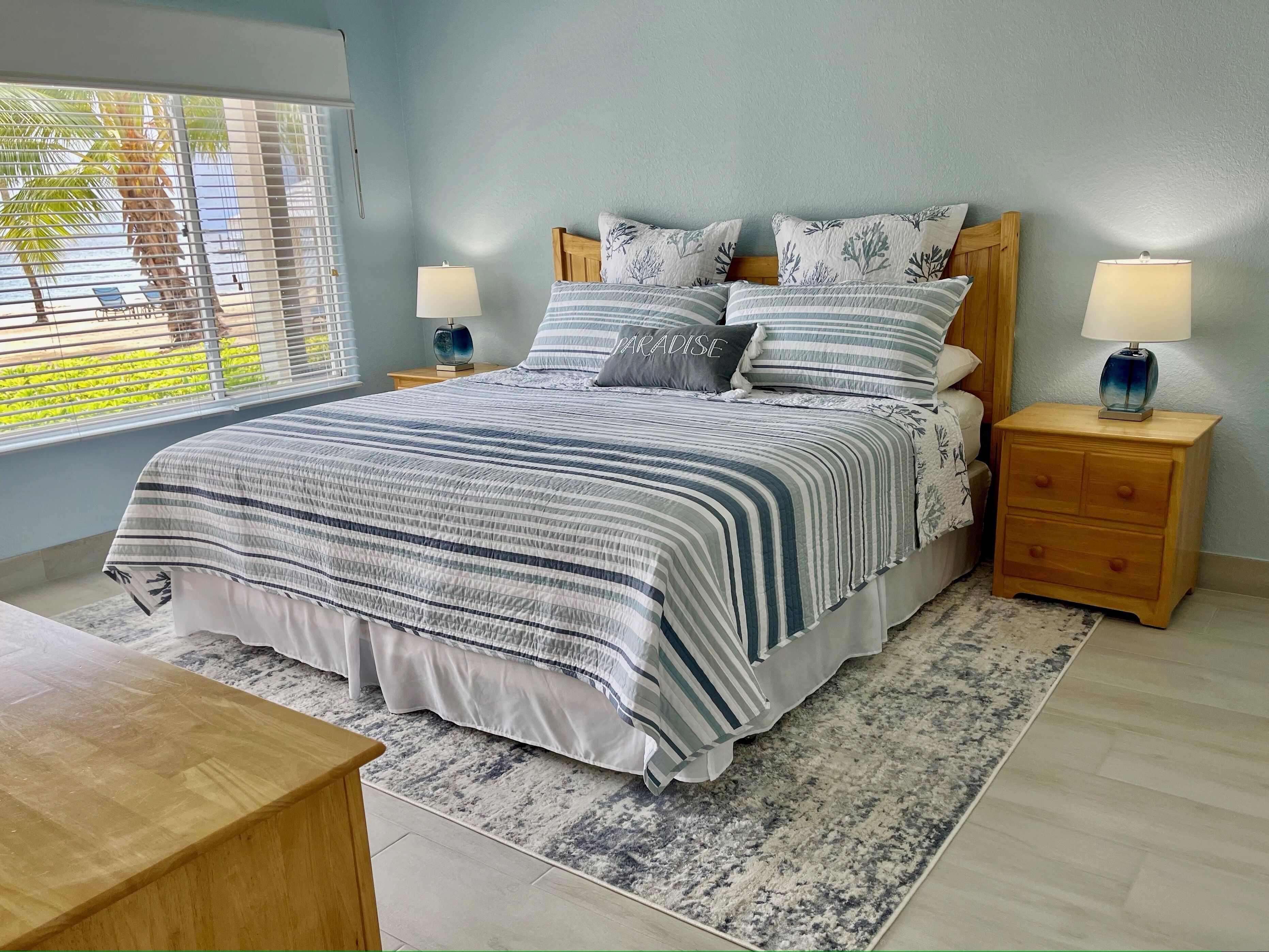 Un dormitorio con una cama grande, dos mesitas de noche con lámparas, una mesita auxiliar de madera, una alfombra estampada y una ventana con persianas.