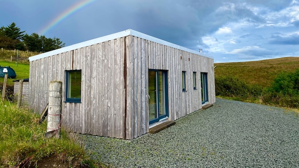 A modern wooden cabin with a gravel driveway and a rainbow in the background. - The Cabin (Dunvegan)