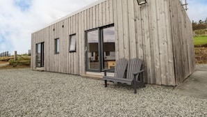 A modern wooden cabin with large sliding glass doors, a small porch with two Adirondack chairs, and a gravel ground. - The Cabin (Dunvegan)