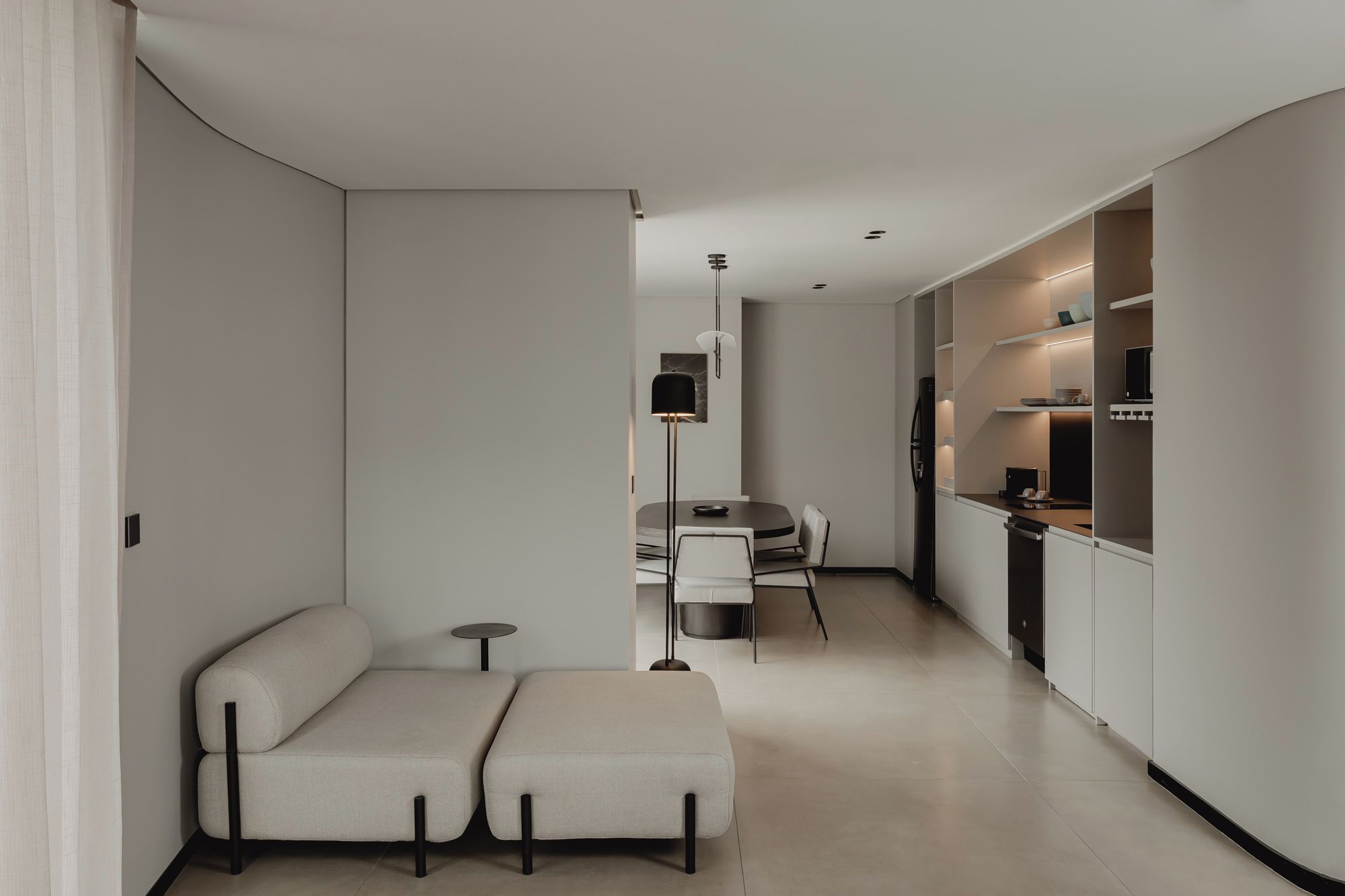 Una sala de estar moderna y minimalista con un sofá y reposapiés, un comedor con mesa y sillas, y una cocina con electrodomésticos integrados y estanterías.