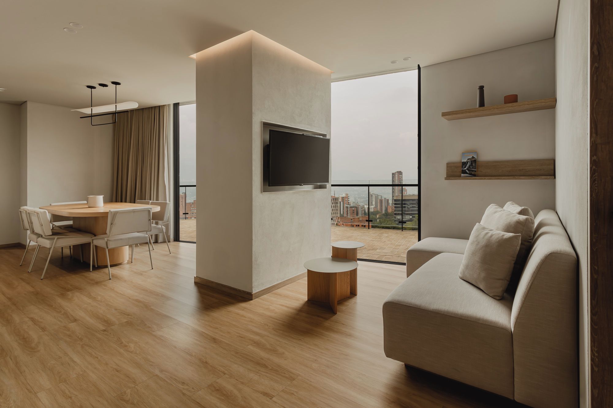 Una sala moderna con un sofá, una mesa de comedor con sillas, un televisor montado en la pared y un balcón con vistas a la ciudad.