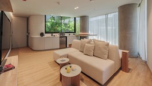 Un soggiorno moderno con un divano beige, due tavolini rotondi e un angolo cottura con una grande finestra che si affaccia sul verde.
