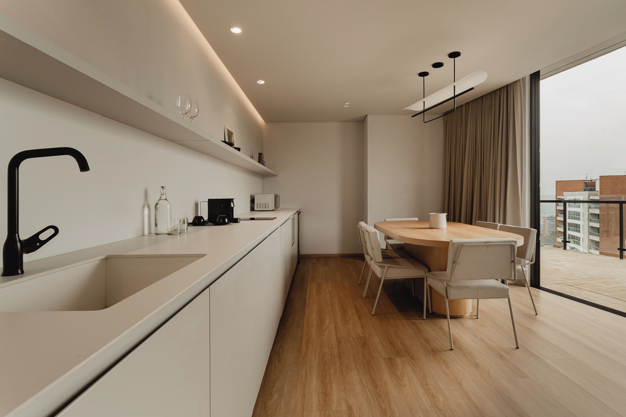 Una cocina moderna con una isla central, una zona de comedor con una mesa redonda y un ventanal con vistas a edificios.