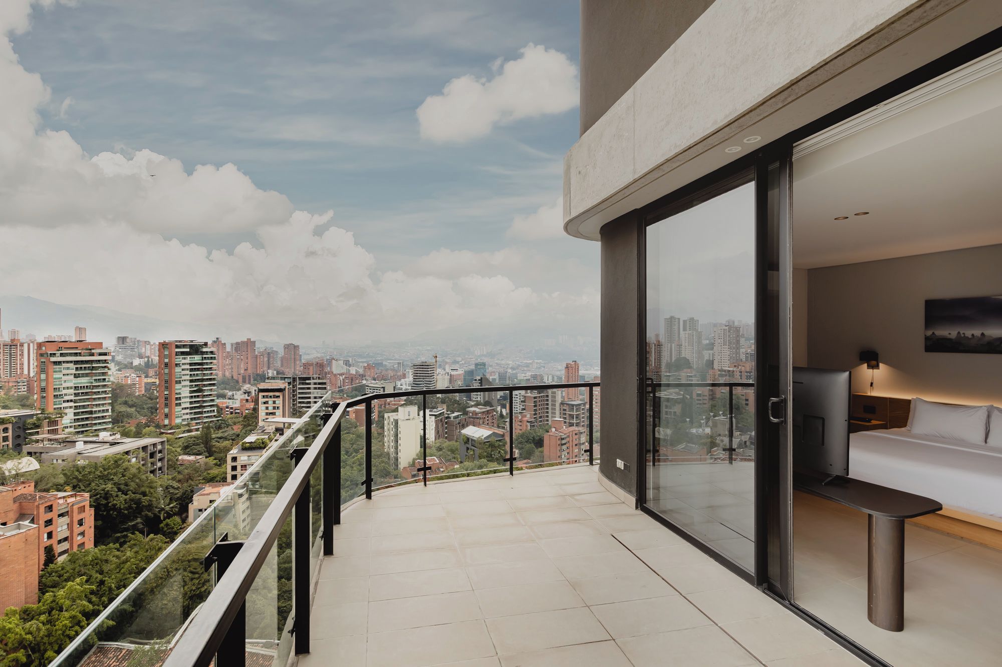 A modern balcony with a city view, a glass door leading to a bedroom with a bed and a TV, and a clear sky.