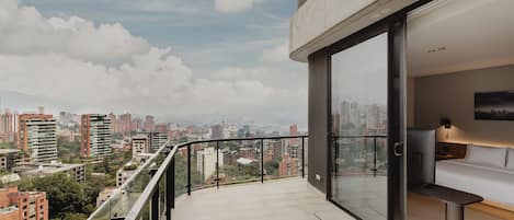 Varanda moderna com vista para a cidade, porta de vidro que dá para um quarto com cama e televisão, e céu limpo.