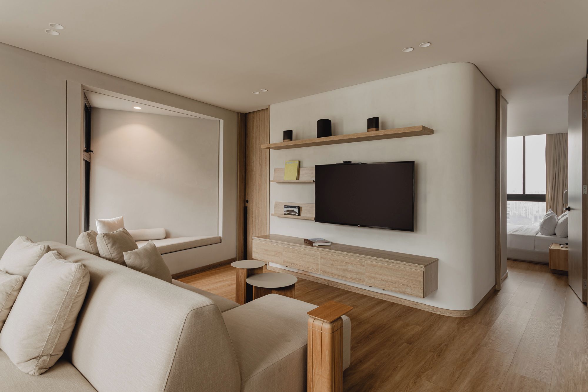 A modern living room with a sofa, a flat-screen TV mounted on a wooden wall, a small round table, and a window with a view.