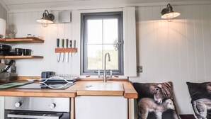 Une cuisine dotée de plans de travail en bois, d’un four encastré, d’un évier et d’une fenêtre offrant une vue agréable. On y trouve deux luminaires fixés au mur et une étagère contenant des ustensiles de cuisine.