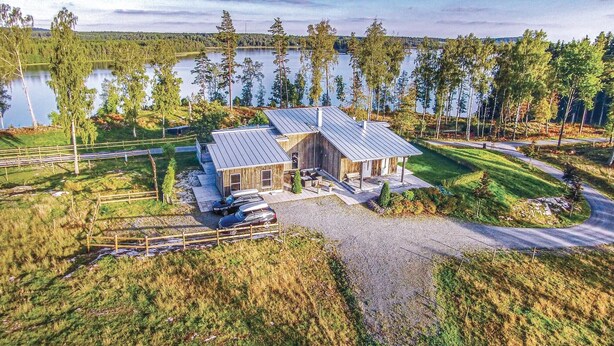 Et moderne hus med metal tag, omgivet af et naturligt landskab med sø og træer.