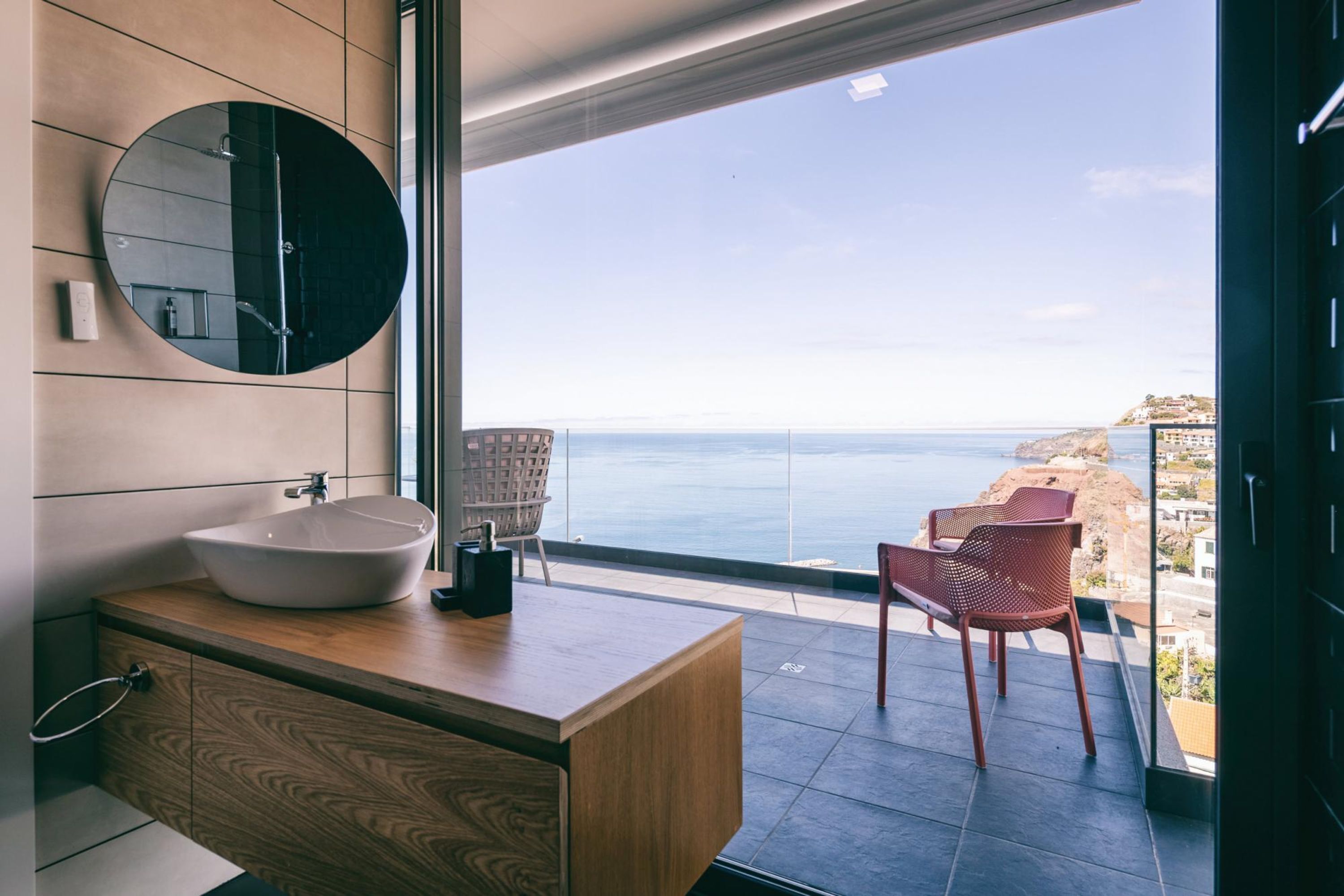 Un baño moderno con un espejo redondo grande, un tocador de madera y vista al mar.