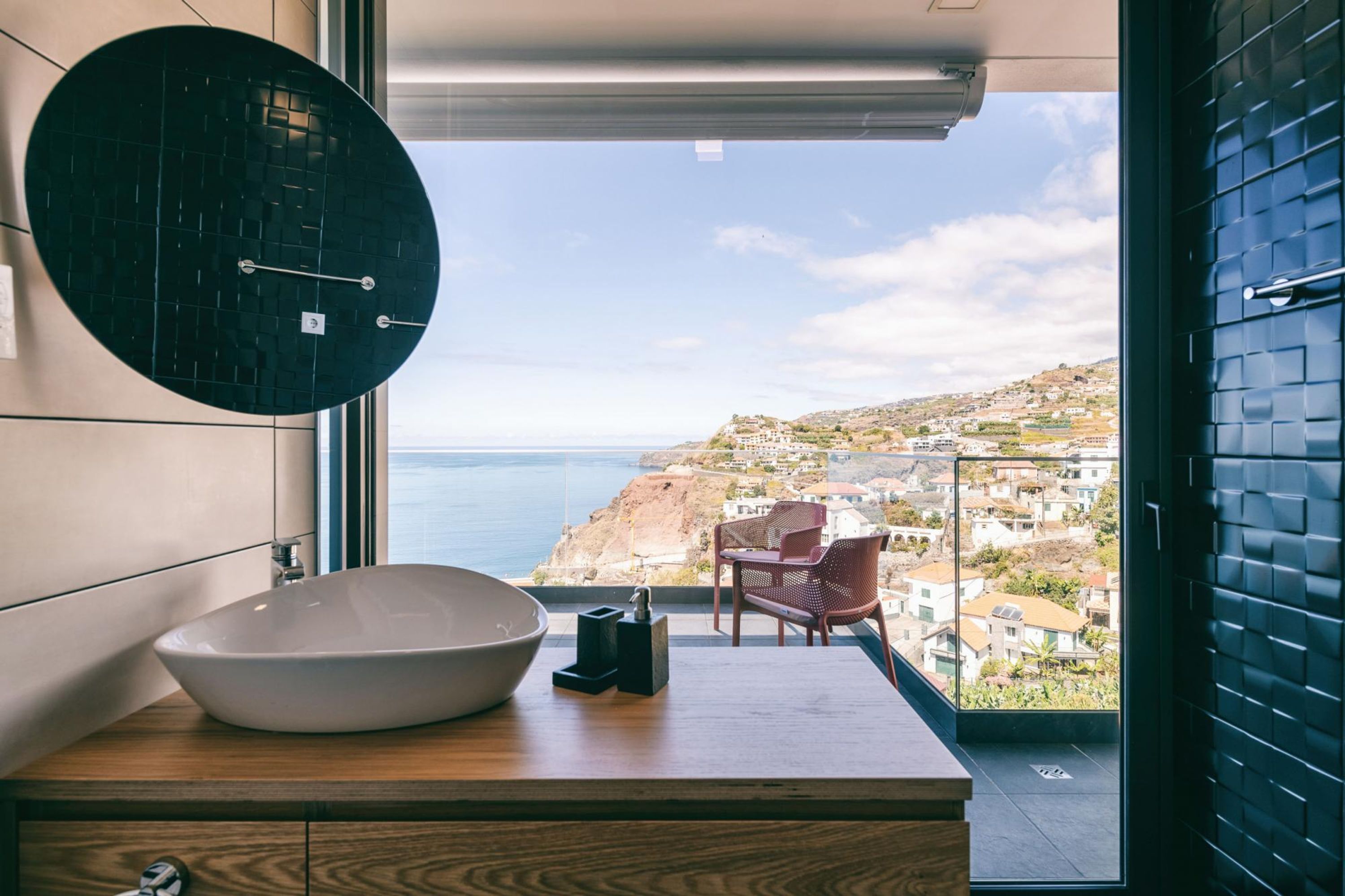 A modern bathroom with a large round mirror, a white sink, and a view of a coastal town through a large window.