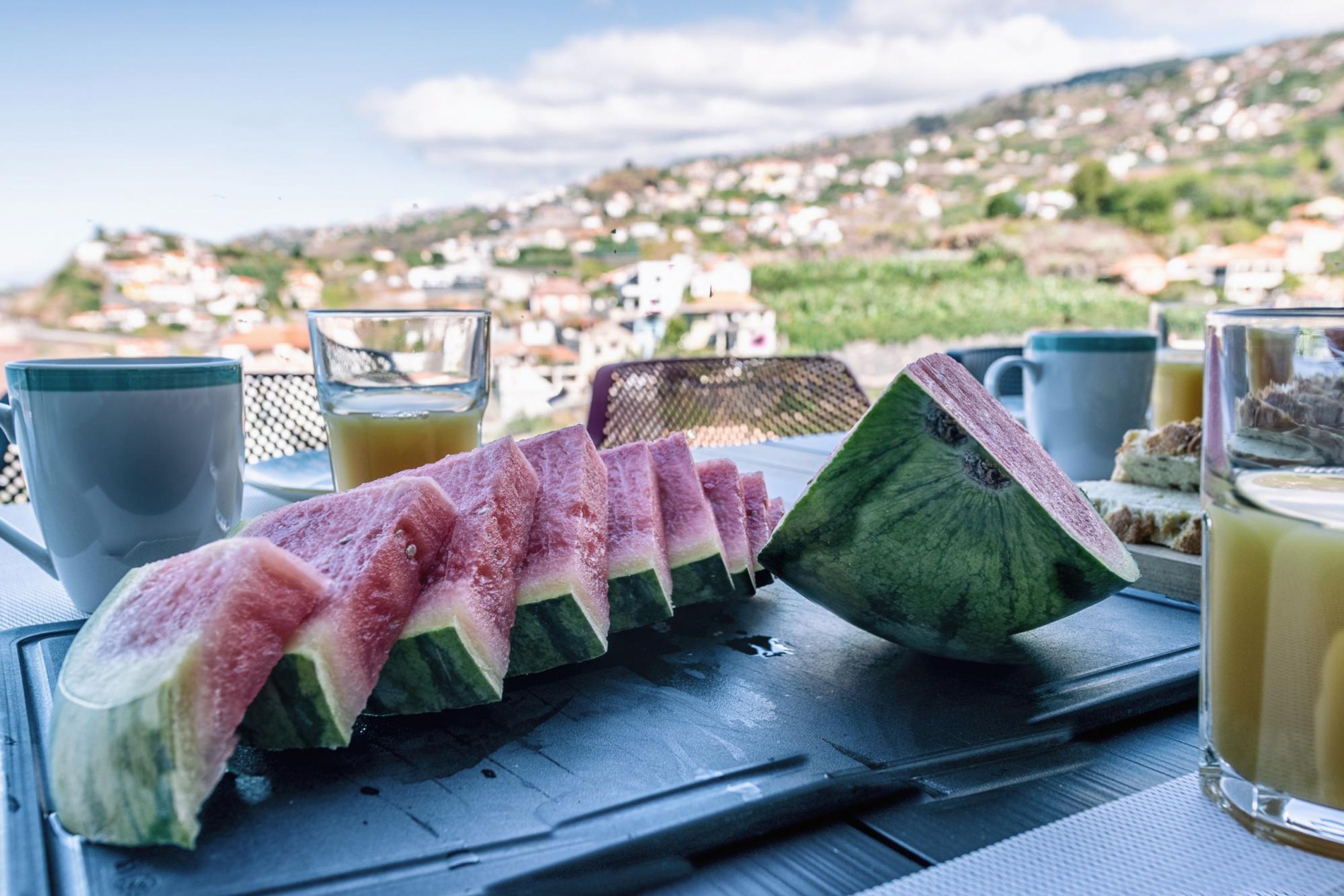 Une table avec des tranches de pastèque, un verre de jus d’orange et une tasse de café, le tout disposé devant un paysage pittoresque d’une colline.