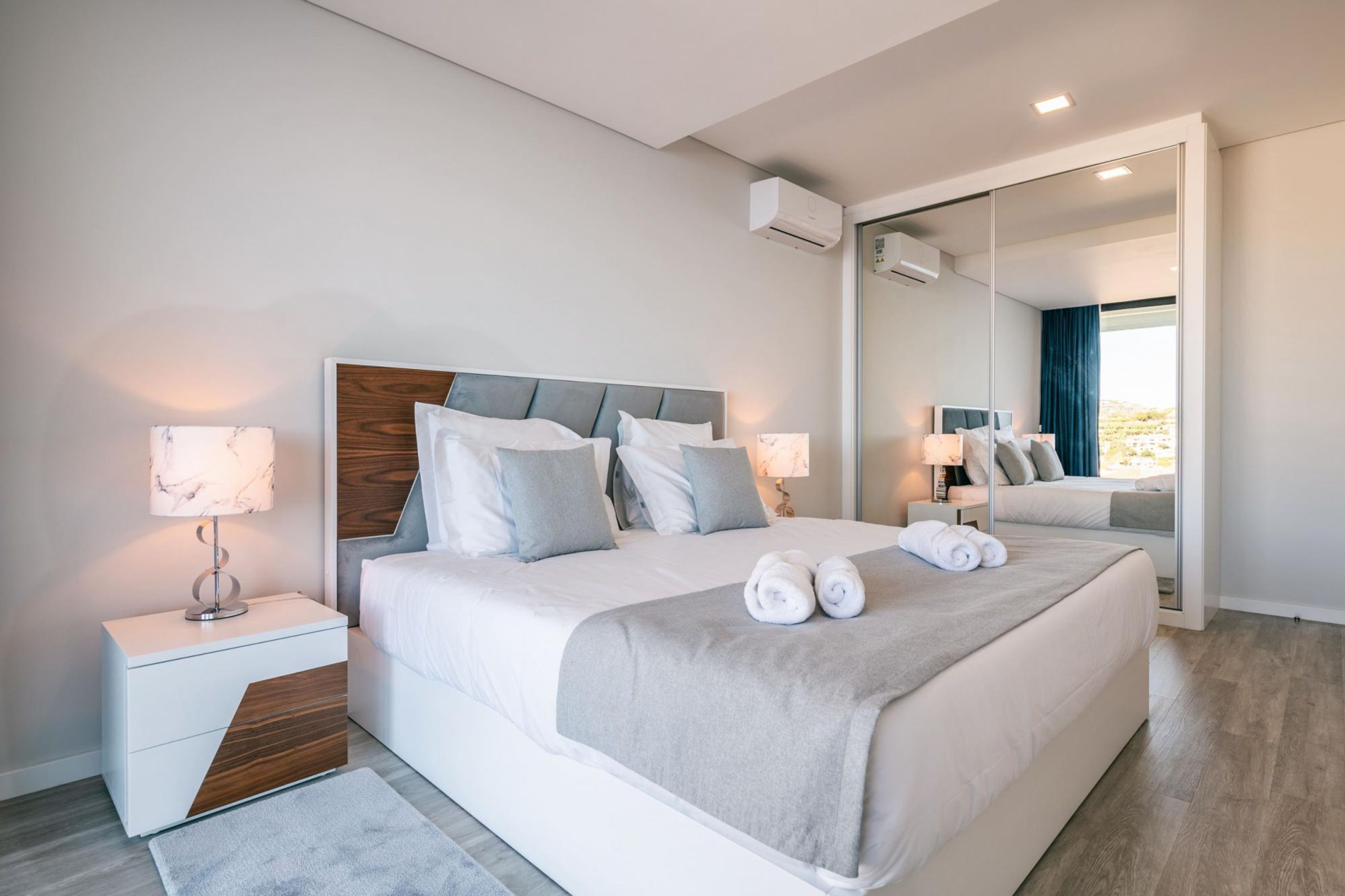 A modern hotel room with a large bed, a bedside table with a lamp, a mirror, and an air conditioning unit.
