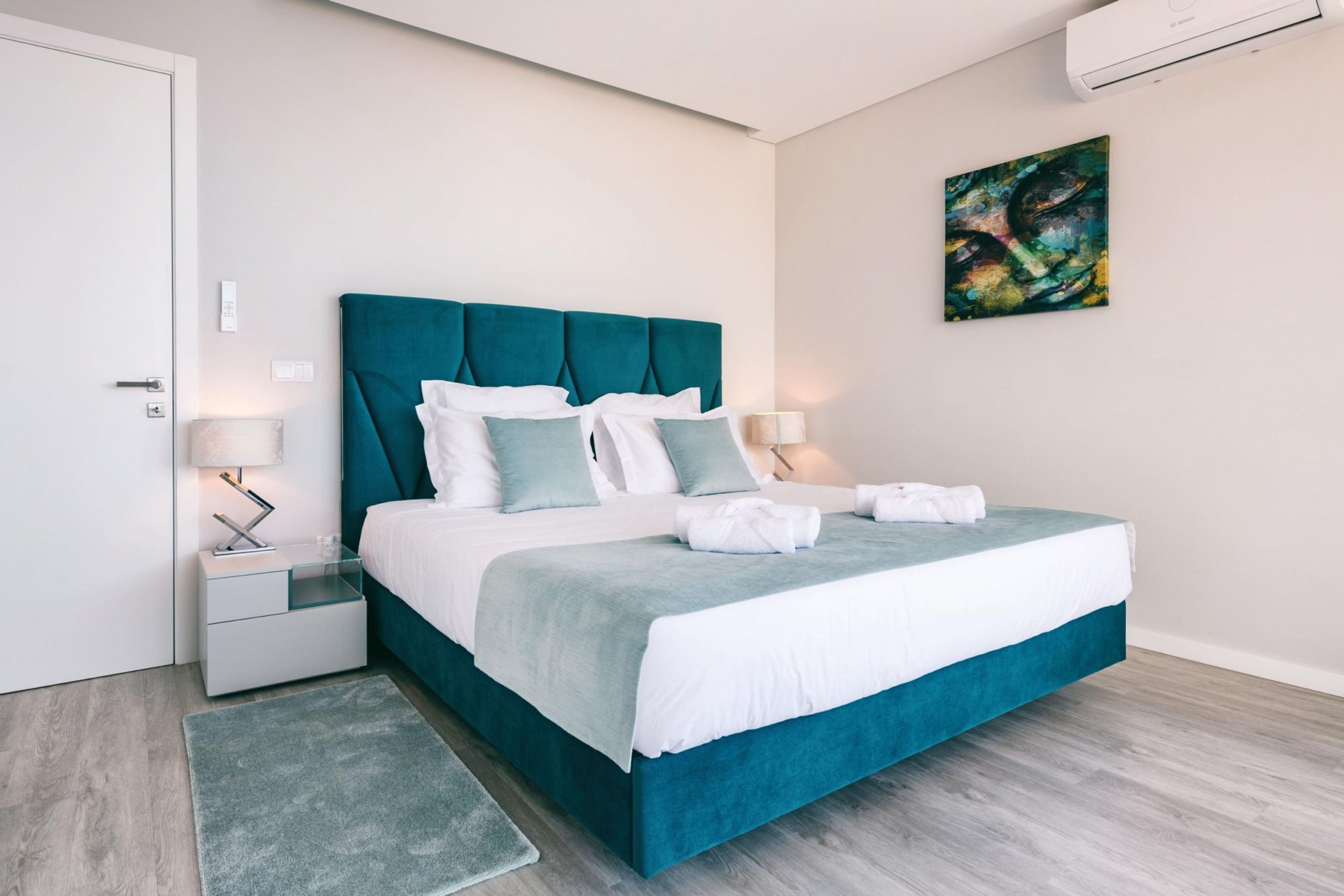 Un dormitorio moderno con un tablero de cama tapizado en color verde azulado, ropa de cama blanca y una alfombra gris.