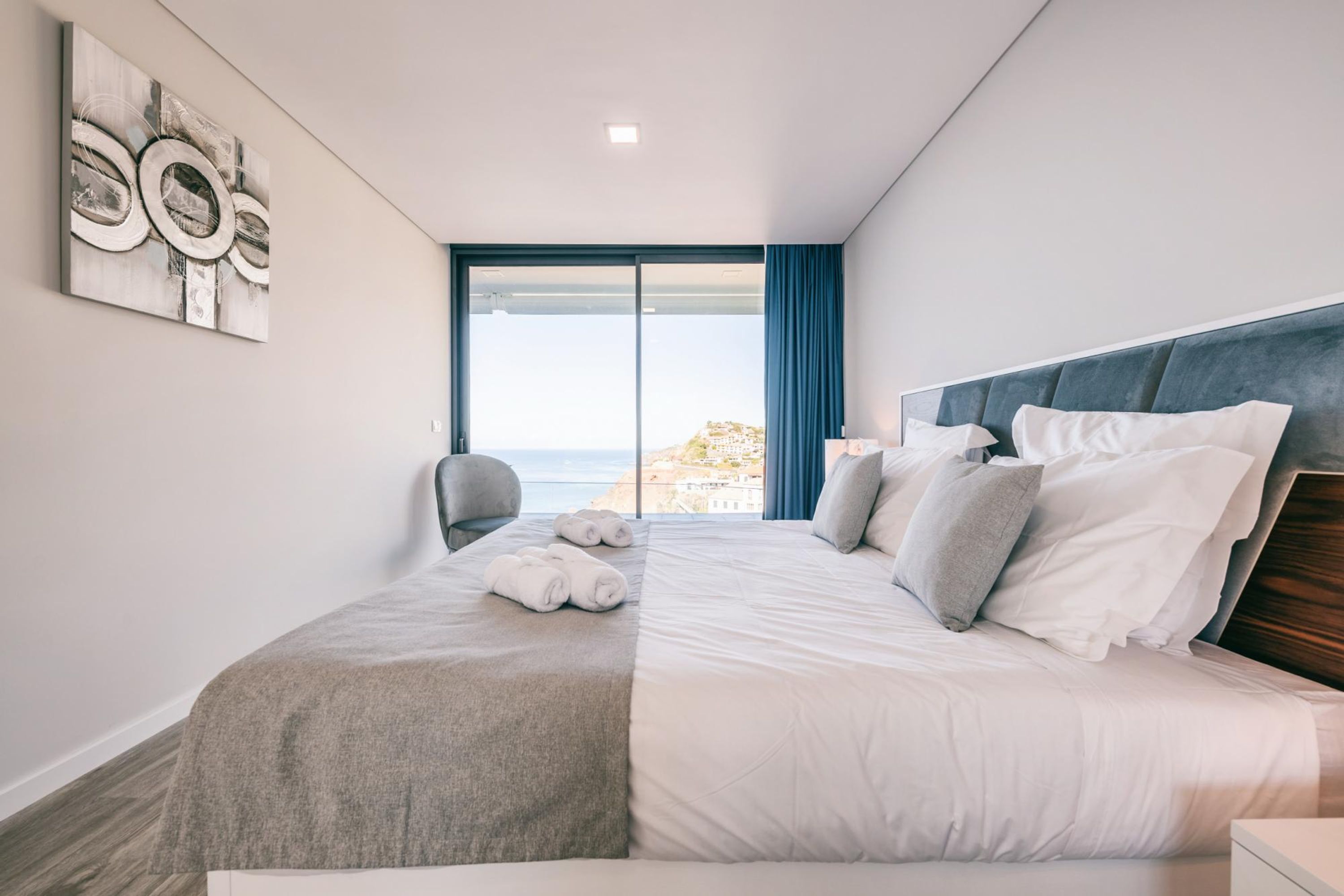 A modern hotel room with a large bed, a view of the sea, and a painting on the wall.