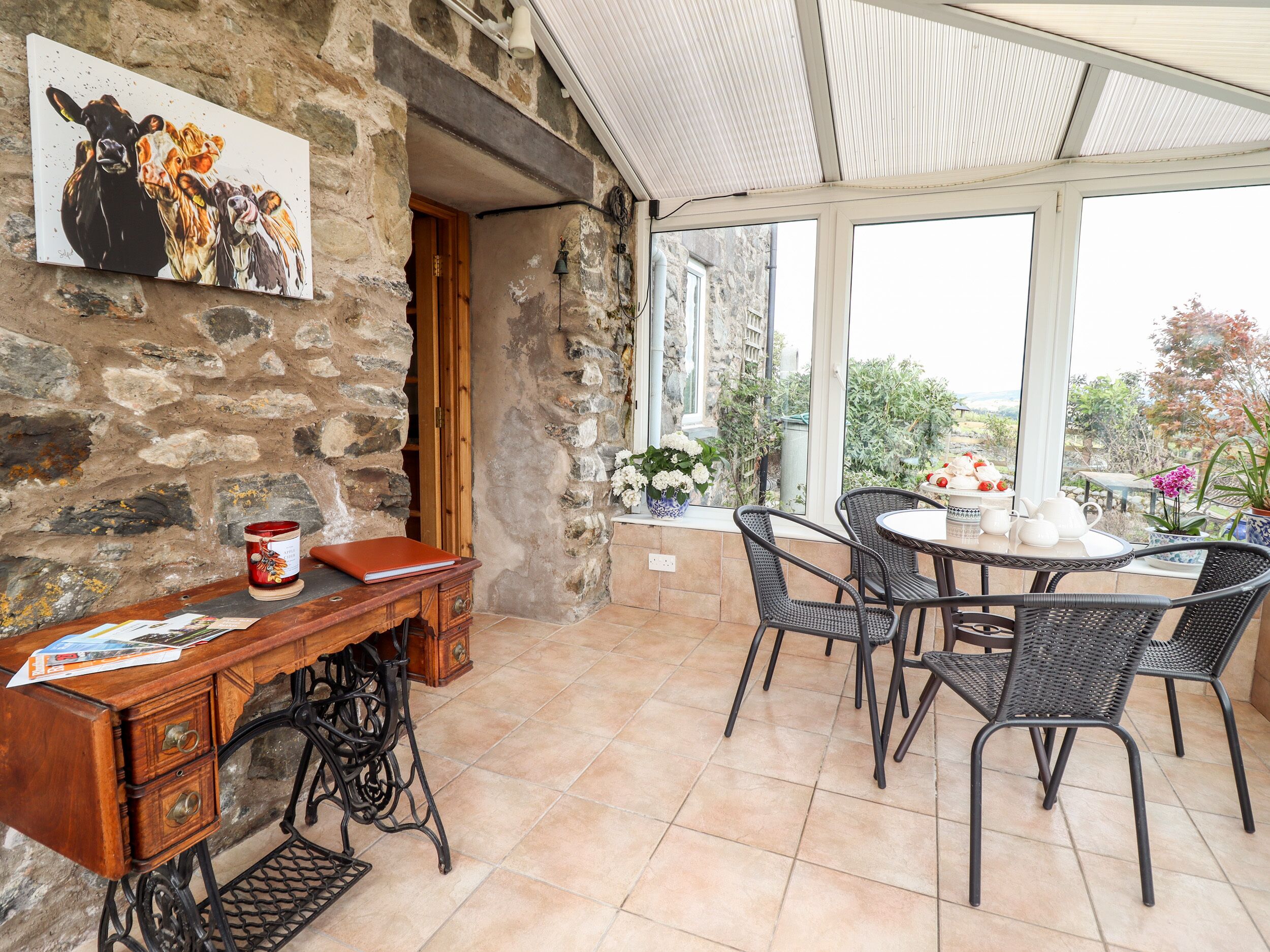 A rustic stone wall with a painting, a wooden desk with a red can, and wicker chairs around a white table.