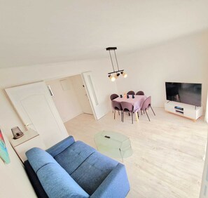 Ein modernes Wohnzimmer mit einem blauen Sofa, einem Glastisch, einem Esstisch mit violetten Stühlen, einem Fernseher und einer Deckenleuchte.