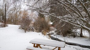 Een houten picknicktafel bedekt met sneeuw, omringd door kale bomen en een besneeuwd pad.