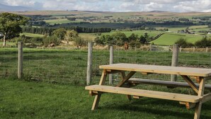 Une table de pique-nique se trouve dans un champ verdoyant, offrant une vue sur des collines ondulantes et une clôture en arrière-plan.