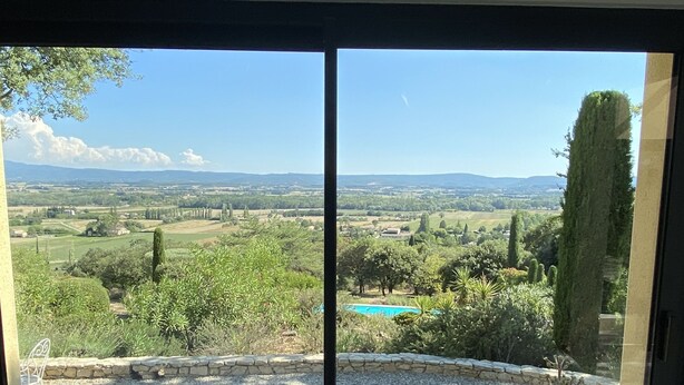 Vista desde una habitación con un gran ventanal corredizo, que muestra una piscina, jardín ajardinado y montañas a lo lejos.