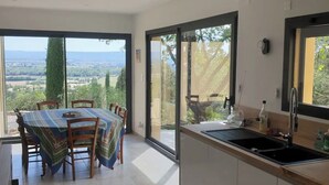 Una cocina moderna con una isla grande, un comedor con mesa y sillas, y vistas al campo a través de amplios ventanales.