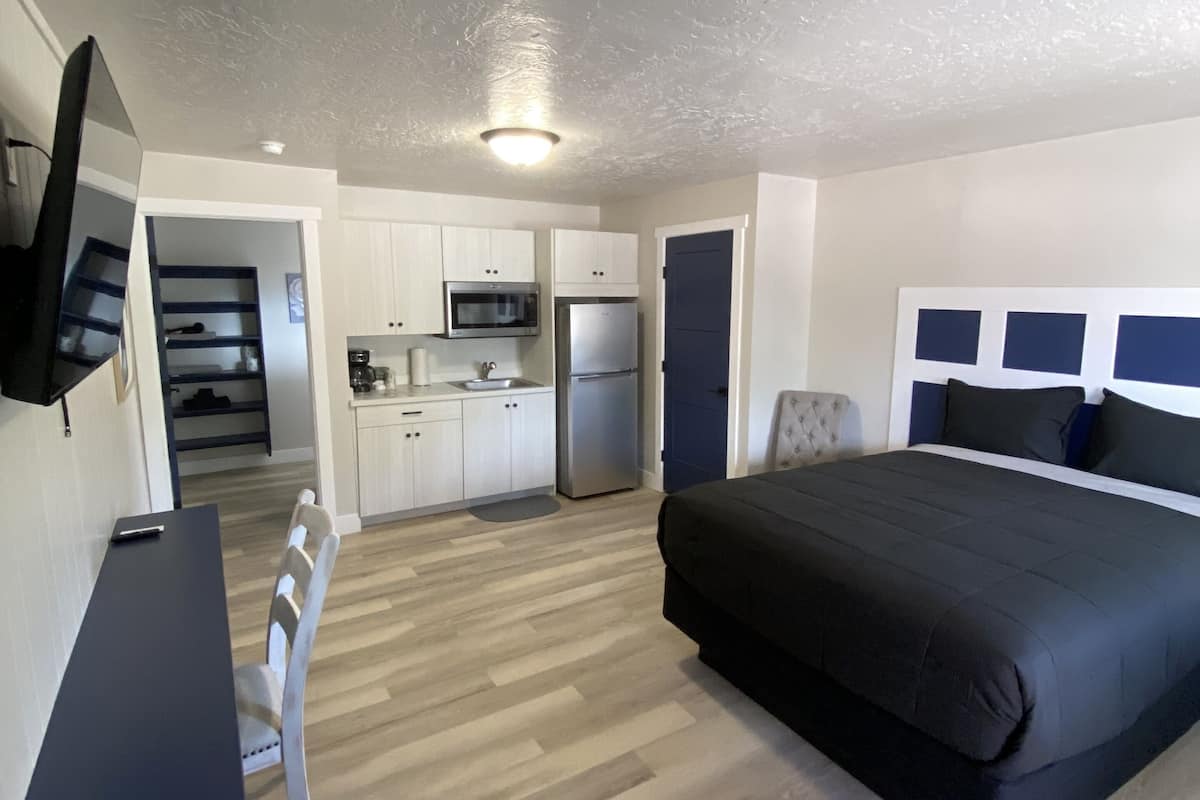 A modern bedroom with a large bed, a desk, and a kitchenette in the background.