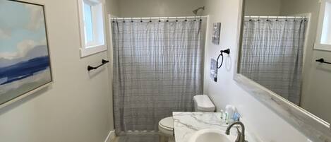 A bathroom with a marble sink, a toilet, a shower curtain, a ceiling fan, and a painting on the wall.