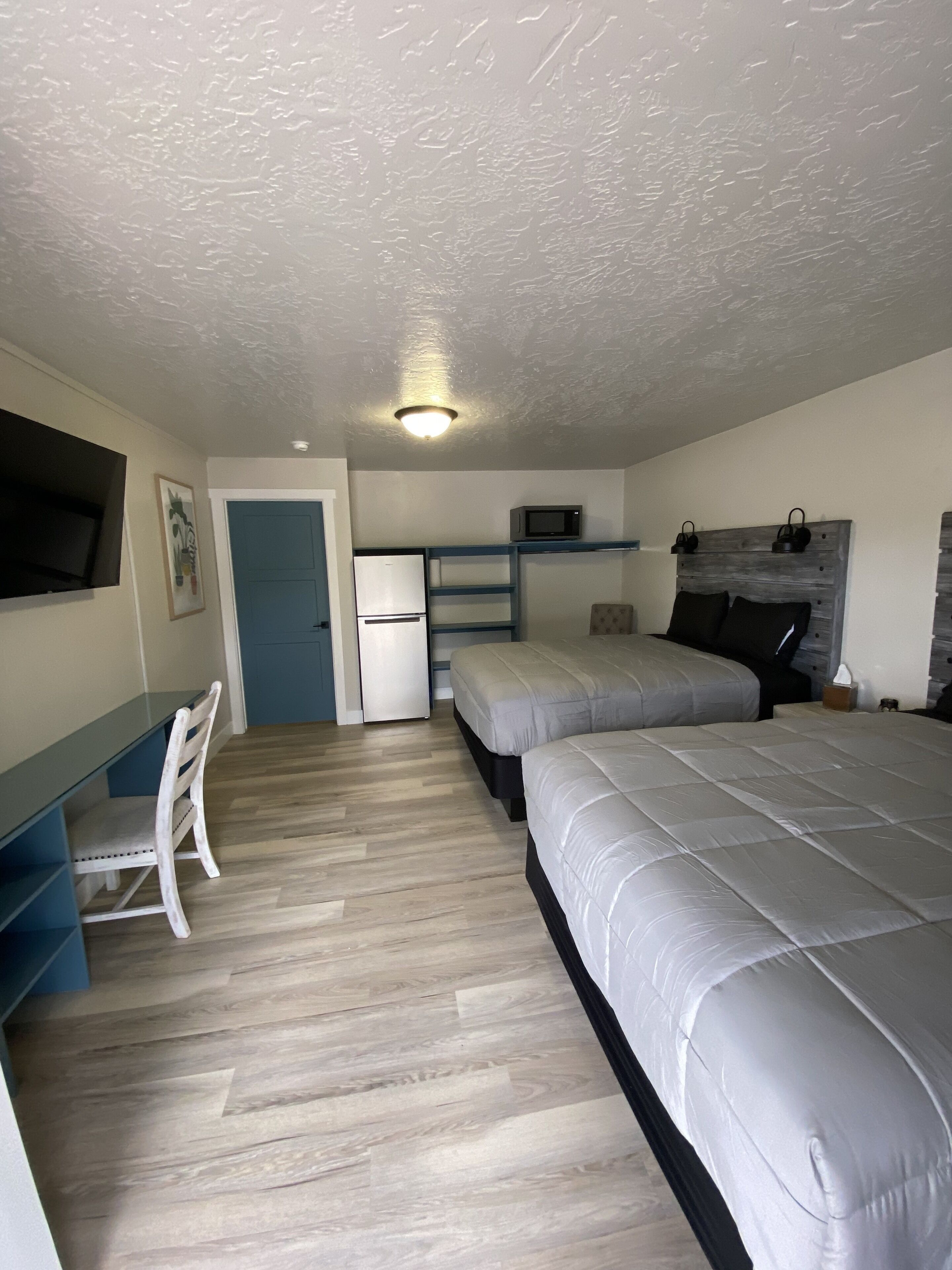 Un dormitorio funcional con dos camas, un escritorio, una silla, un refrigerador pequeño, un microondas y una televisión instalada en la pared.