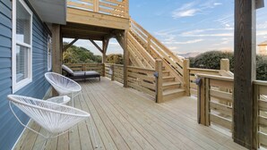 A wooden deck with a staircase leading to a covered area, two white wire mesh chairs, and a bench. - Outer Banks Haven: Pet-Friendly, Walk-to-Beach, EV Charging, Updated Kitchen (Kitty Hawk)
