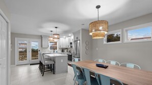 A modern kitchen with white cabinets, a central island, and a dining area with blue chairs. - Outer Banks Haven: Pet-Friendly, Walk-to-Beach, EV Charging, Updated Kitchen (Kitty Hawk)