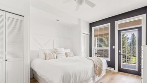 A bedroom with a large bed, white walls, a ceiling fan, and a view of greenery through the windows. - Fairmont Oasis | Luxury | Views | Bright | Nature (Fairmont Hot Springs)