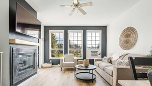 A modern living room with a fireplace, a sofa, a coffee table, and a view of the outdoors through large windows. - Fairmont Oasis | Luxury | Views | Bright | Nature (Fairmont Hot Springs)