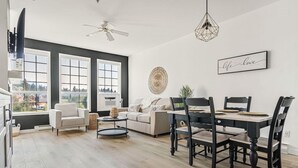 A spacious living room with a dining area, large windows, a ceiling fan, and a wall-mounted artwork. - Fairmont Oasis | Luxury | Views | Bright | Nature (Fairmont Hot Springs)