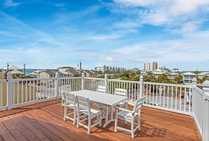 Une terrasse en bois avec une table et des chaises blanches, entourée d’une clôture blanche, donnant sur un quartier résidentiel avec des maisons et un ciel dégagé.