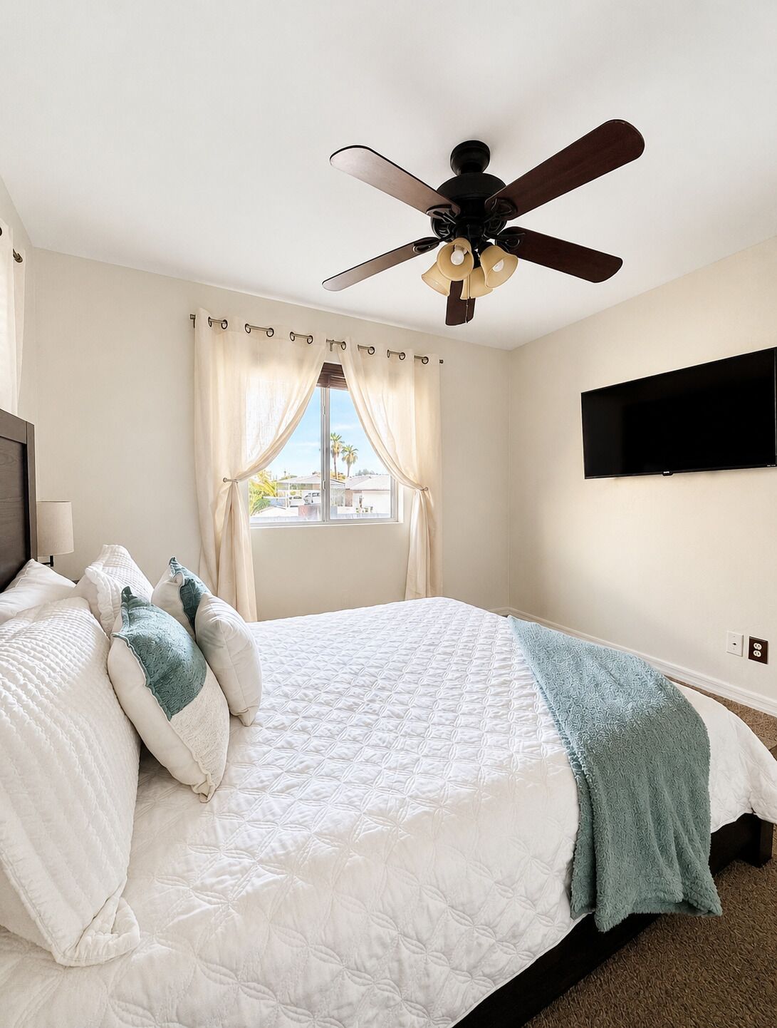 A bedroom with a bed, a ceiling fan, a window with curtains, a TV mounted on the wall, and a view of the outside.