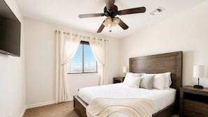 Un dormitorio con una cama, ventilador de techo, mesita de noche, lámpara y ventana con cortinas.