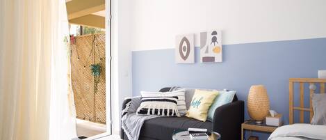 A modern living room with a sofa, a small round table, and a bed. There is a sliding door leading to an outdoor area.