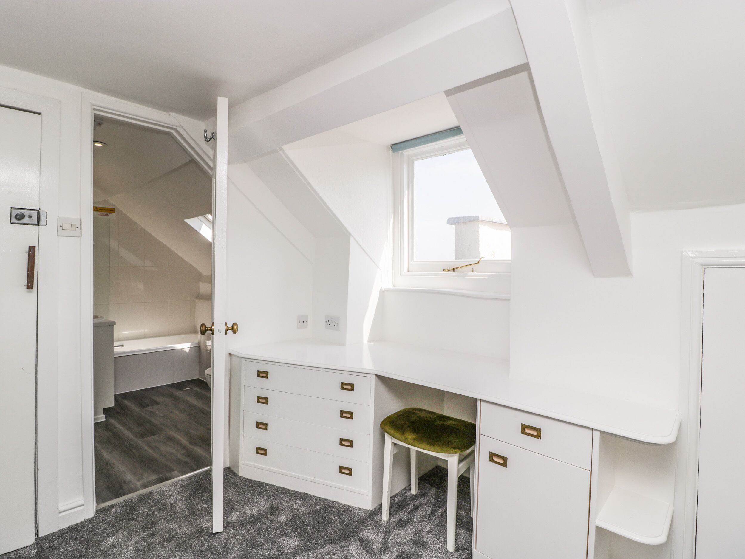 A white room with a sloped ceiling, a small window, and a built-in desk with drawers.